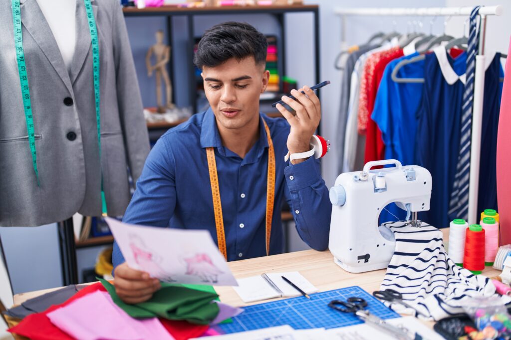 Young hispanic man tailor talking on smartphone looking clothing design at clothing factory